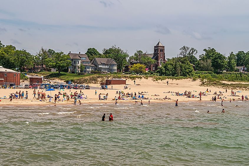Saint Basil Catholic Church behind a bustling beach on Lake Michigan in South Haven, Michigan, USA. People playing and lush green foliage in the background.