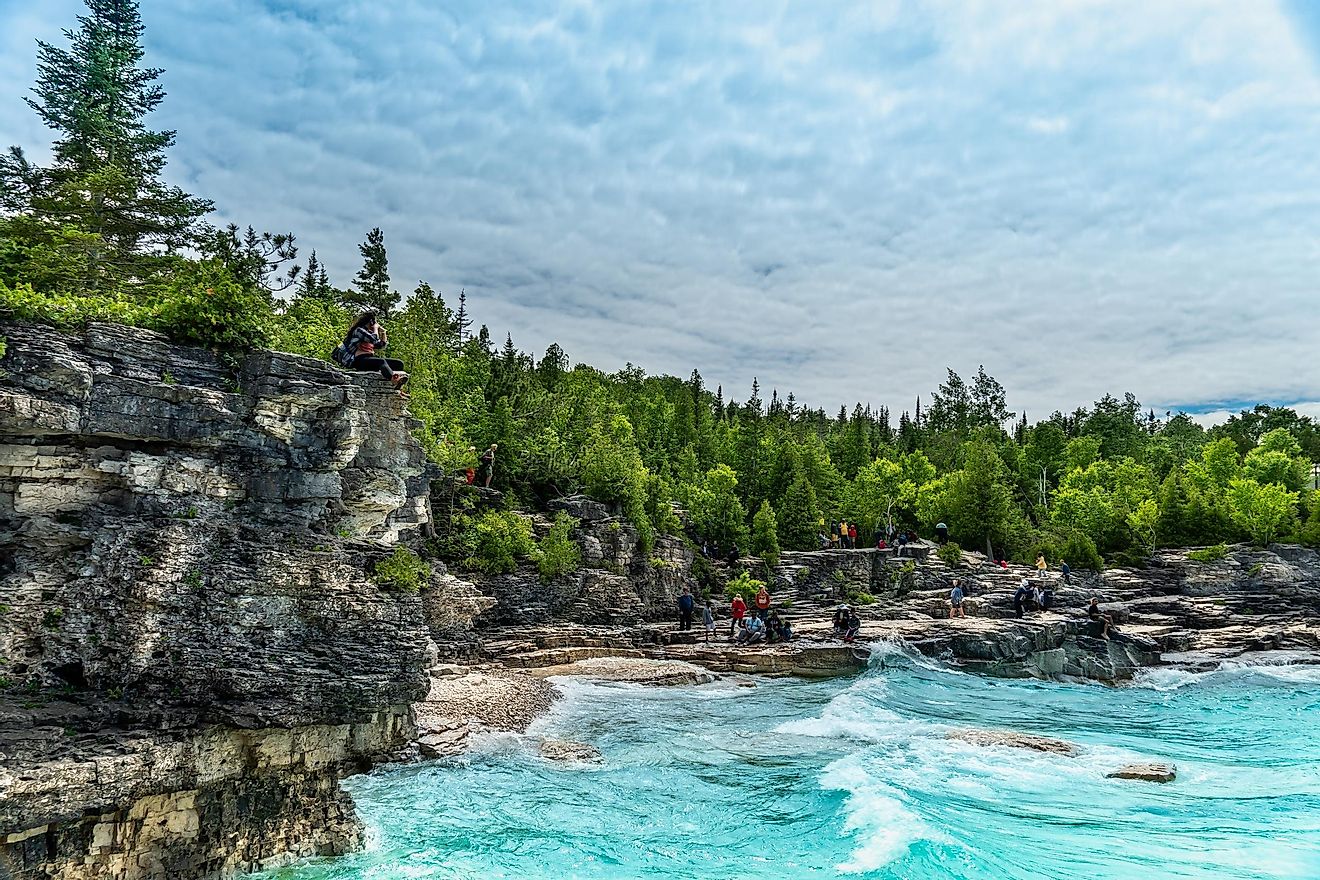 Backpackers taking a break on limestone cliffs before the rolling waves of Georgian Bay. A dense coniferous forest and overhead clouds form the backdrop. Image by Golden Shrimp via Shutterstock.com