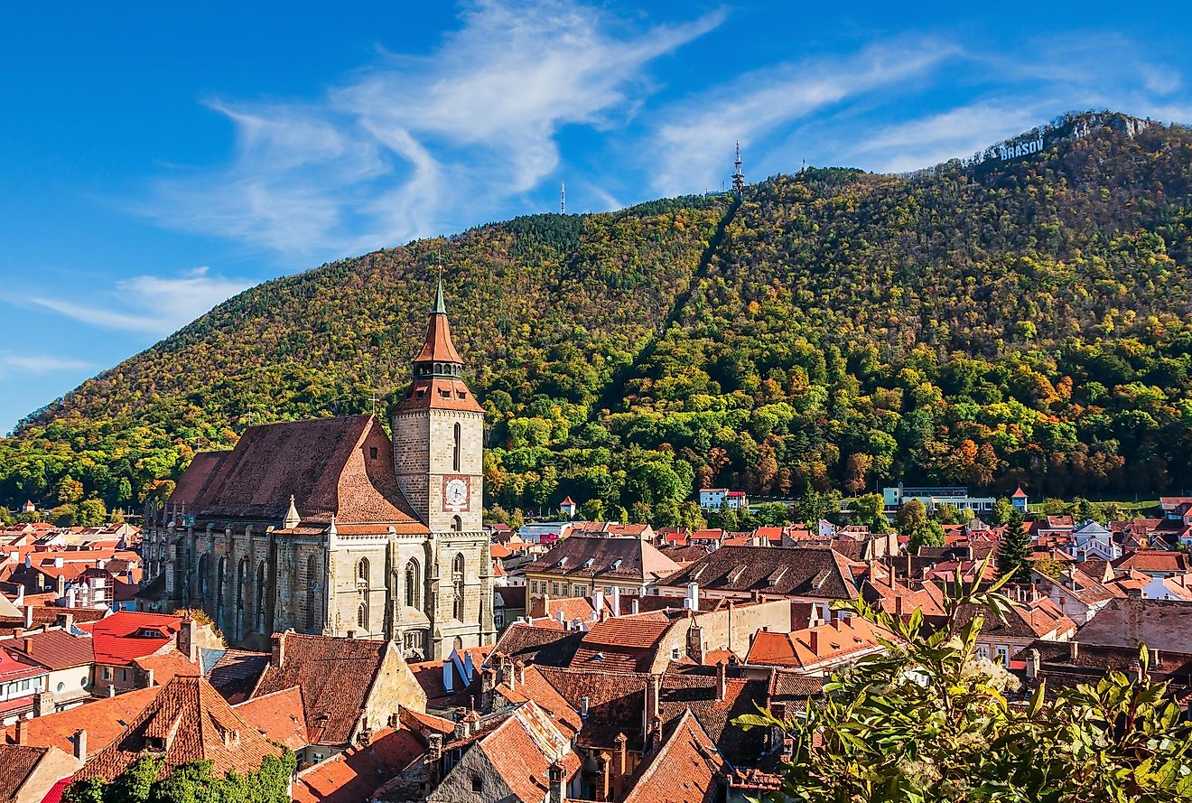 A large green mountain with a "Brasov" Hollywood-style sign stands behind the red-roofed old town, and dominant cathedral.