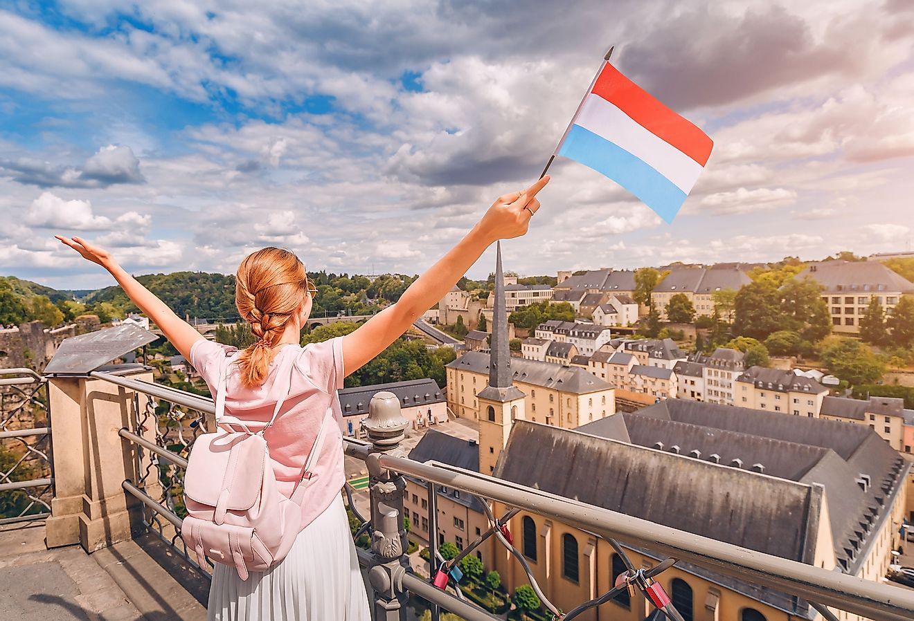 A happy traveler girl holds the flag of Luxembourg and admires the Grund area from the observation deck.