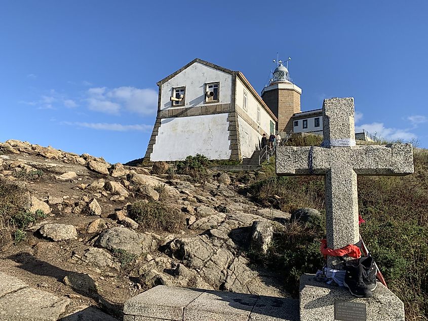 A cross planted on a rocky outcrop. A small lighthouse can be seen in the background.