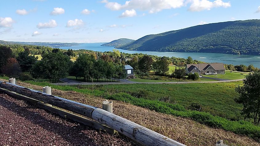 Canandaigua Lake, New York, looking north on a partly cloudy summer day. Rolling hills and trees with a wooden fence and gravel in the foreground.