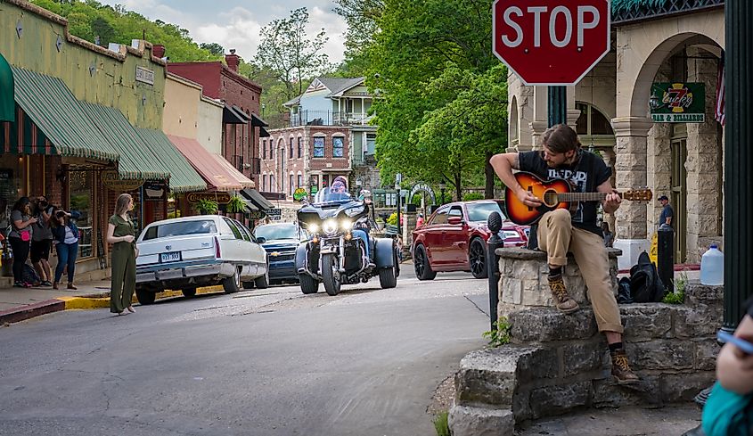 Motorcycle riders cruising downtown Eureka Springs, Arkansas, with a man playing guitar at a stop sign.