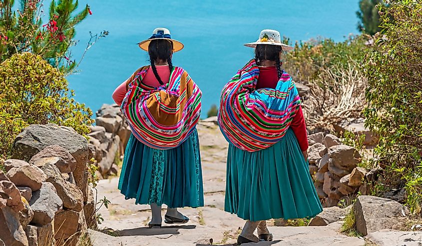 Two indigenous Quechua women in traditional clothes walking down the path to the harbor of Isla Taquile with the Titicaca Lake in the background, Peru
