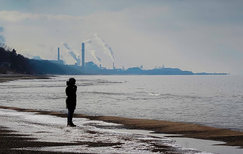 A woman stands on a beach on a late, dim afternoon, with a string of steel mills capturing the skyline. 