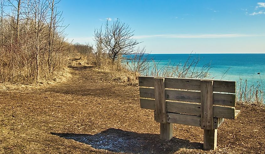 On a sunny day in early Spring, an empty wooden bench faces a peaceful Lake Michigan from atop a bluff at Lion's Den Gorge, near Grafton
