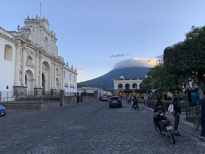 A cobblestone street with an old colonial church on the left, a park on the right, and a cloud covered volcano in the distance 
