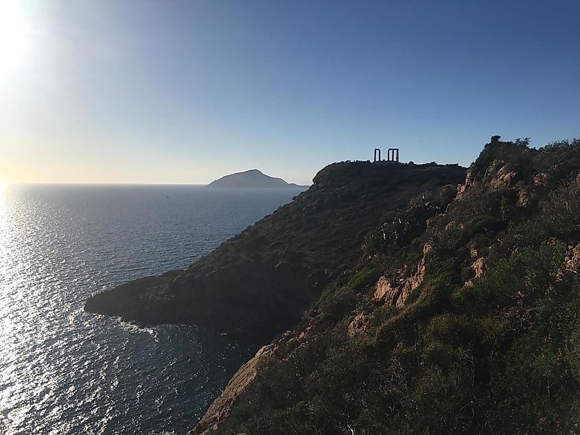 A distant Poseidon's Temple overlooks the sea and the nearly setting sun. Steep, shrub-lined cliffs surround the ancient wonder, and an uninhabited island can be seen just offshore. 