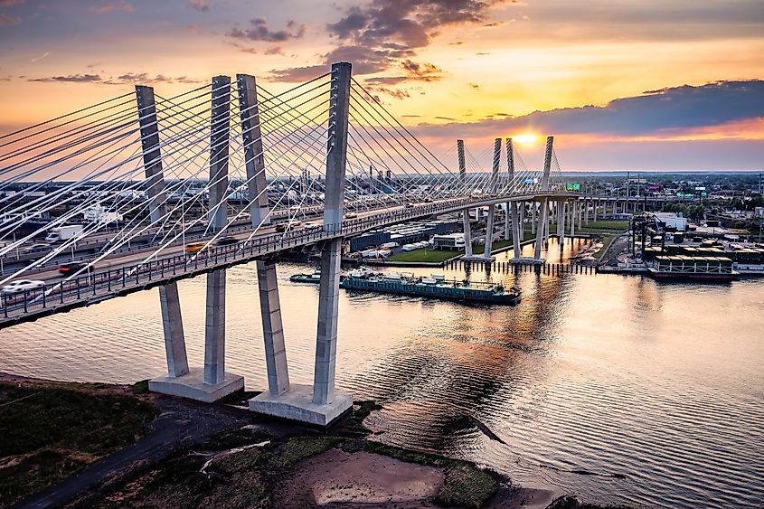 Aerial view of the New Goethals Bridge at sunset, spanning the Arthur Kill strait between Elizabeth, New Jersey, and Staten Island, New York. A container ship navigates under the bridge.