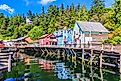 The waterside stores in Ketchikan, Alaska. Colorful wooden buildings on stilts line a scenic waterfront, reflecting in the clear water. Lush green forest and a blue sky complete the vibrant, tranquil scene.