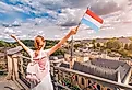 A happy traveler girl holds the flag of Luxembourg and admires the Grund area from the observation deck.