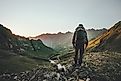 A backpacker stands above a green mountainous valley, looking back towards the setting sun.