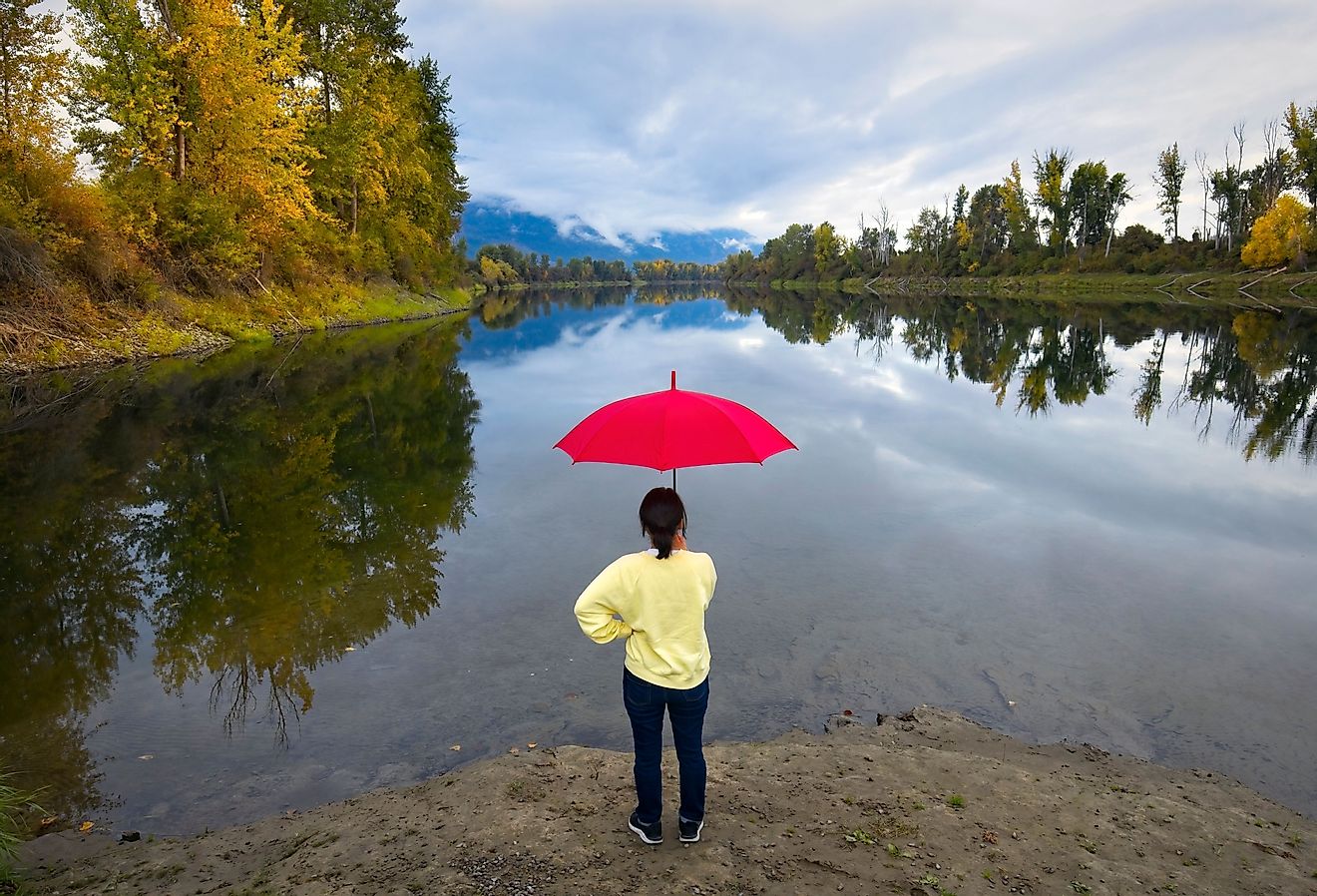 A woman holding a red umbrella stands on the shores by the calm Kootenai River near Bonners Ferry, Idaho.