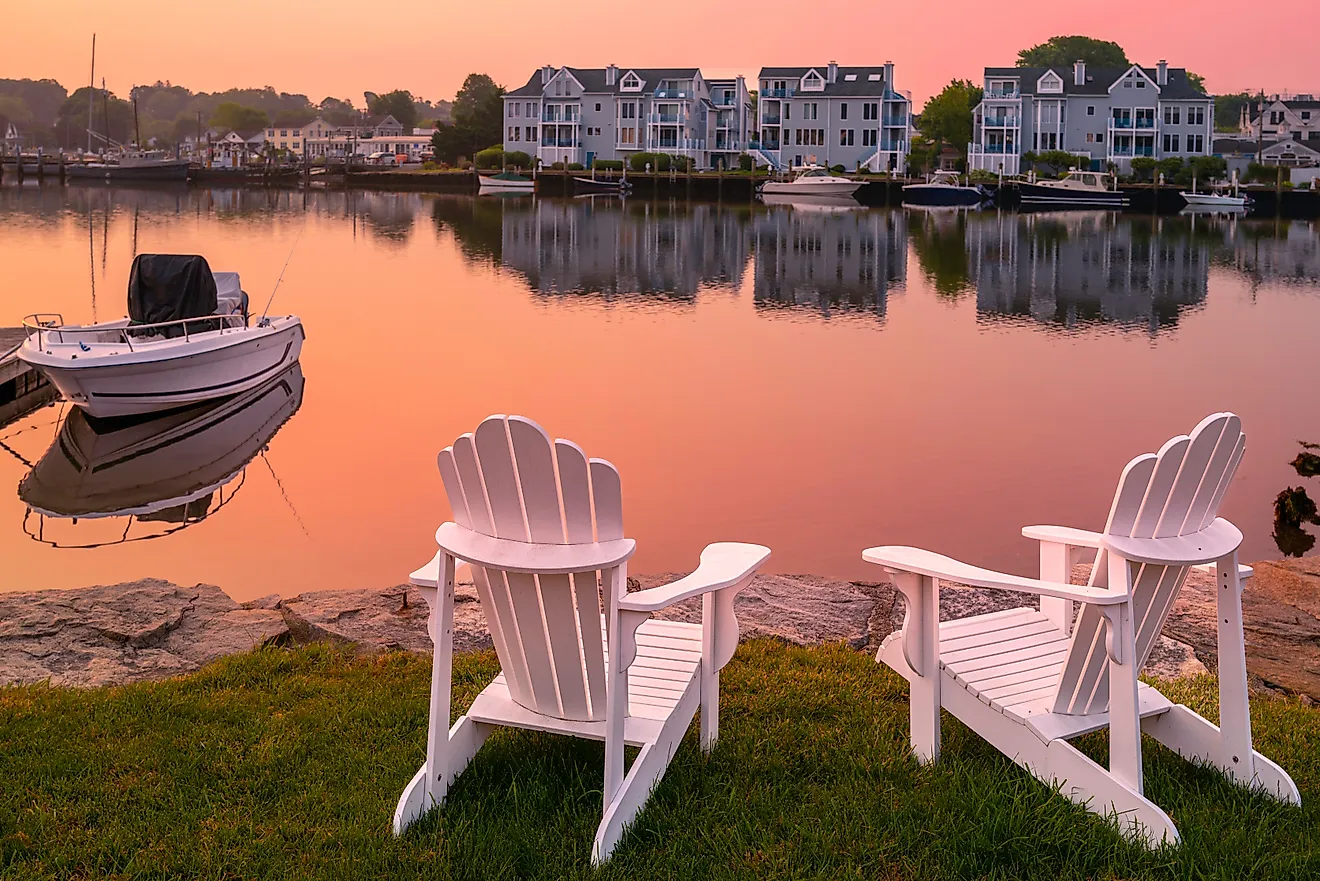 Sunrise, two white Adirondack chairs on the beach, a moored boat, and wildfire smoke over the Mystic River marina village in Connecticut.