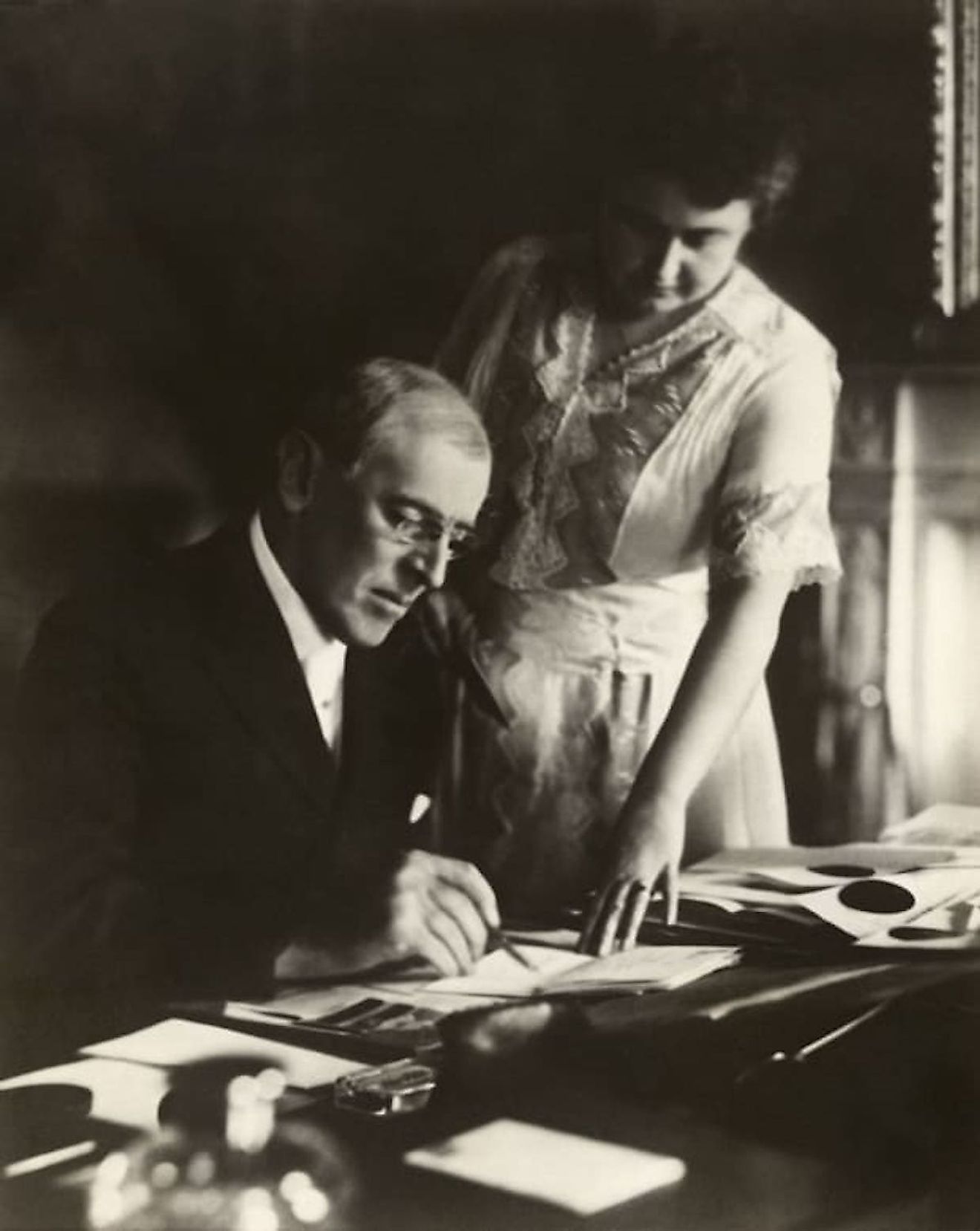 Woodrow Wilson, seated at desk with his wife, Edith Bolling Galt, standing at his side. First posed picture after Mr. Wilson's illness, White House, June 1920. Mrs. Wilson holds a document while the President adds his signature. Image credit: Harris &amp;