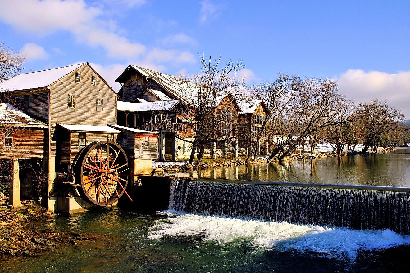 The Old Mill in Pigeon Forge, Tennessee, surrounded by snow, with a picturesque waterwheel and the Smoky Mountains in the background.