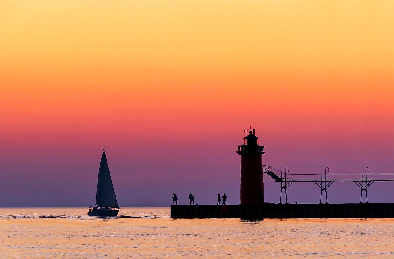 A vividly colorful twilight sky silhouettes a sailboat, people, and the lighthouse at South Haven, Michigan, on Lake Michigan.