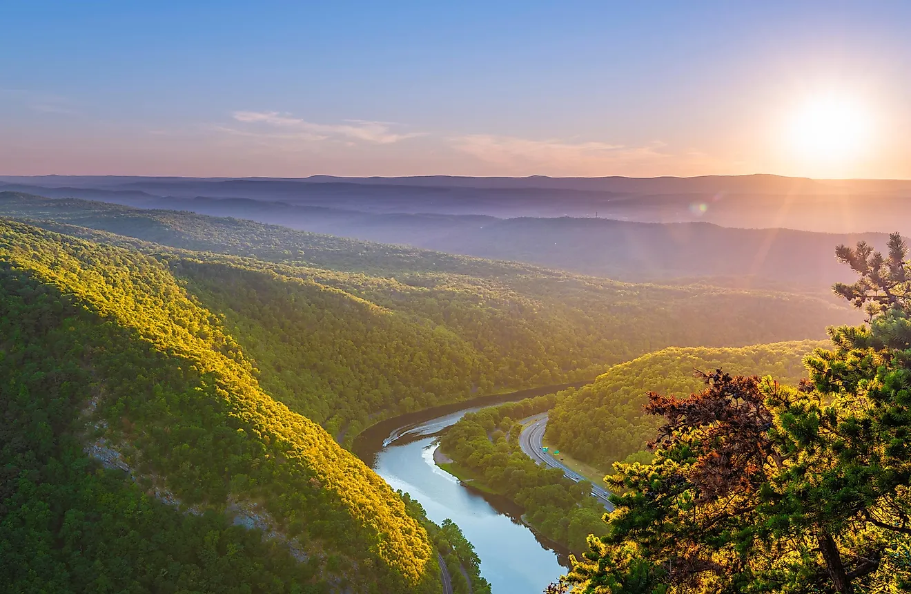 Delaware Water Gap viewed at sunset from Mount Tammany located in New Jersey. Sunset over a lush, green valley with a winding river, surrounded by rolling hills. The sky is clear with warm hues, creating a peaceful atmosphere.