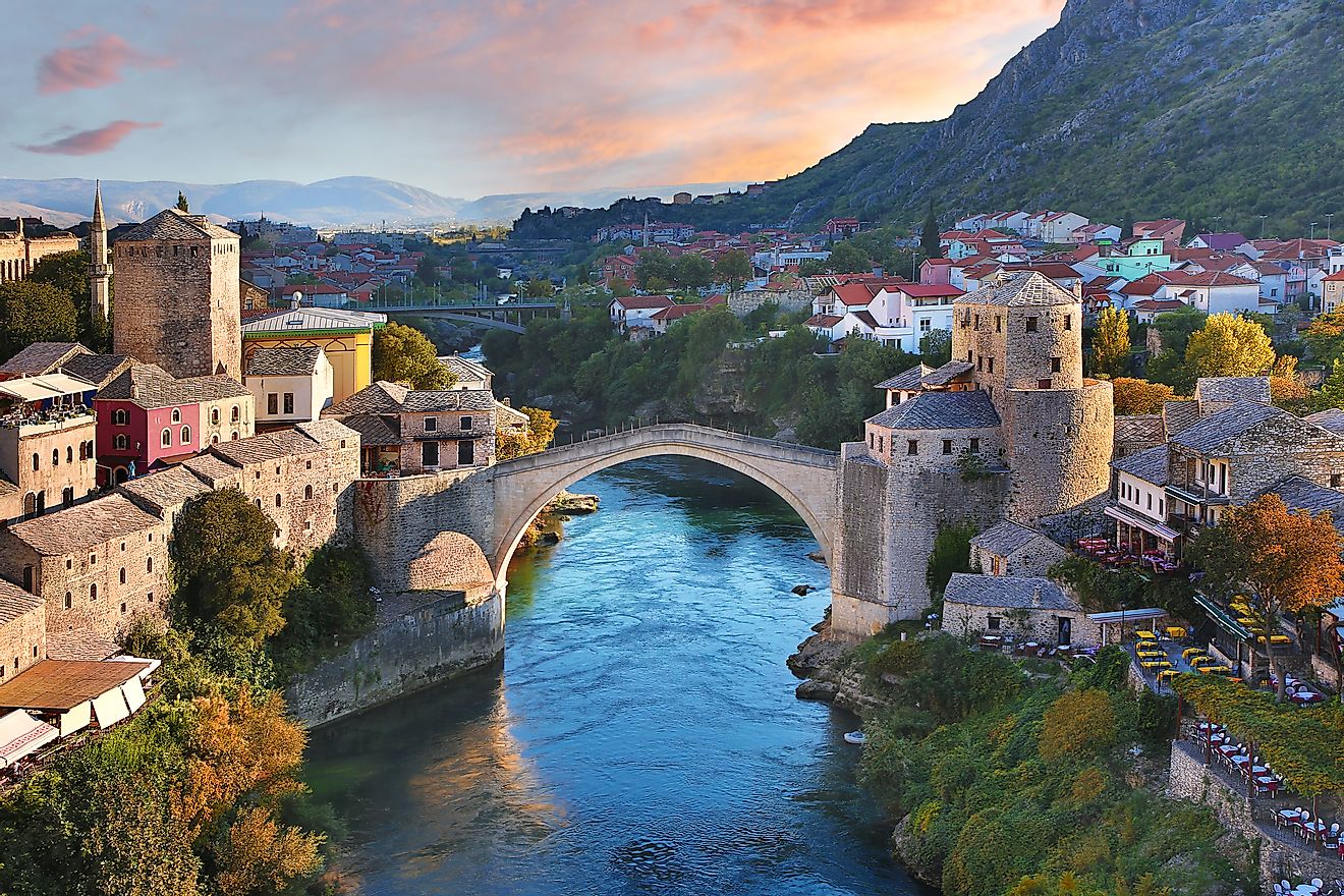 A pre-sunset shot of Mostar's iconic, single arch stone bridge (Stari Most) spanning the emerald blue Neretva River. The antiquitous Old Town encroaches on both banks.