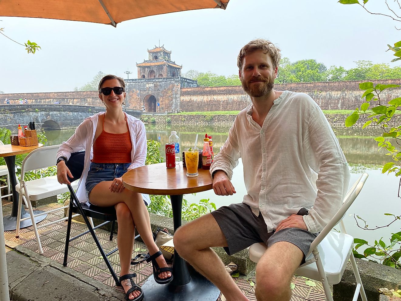 A couple poses for a picture at a café table. An ancient Vietnamese fortress and moat can be seen in the background.
