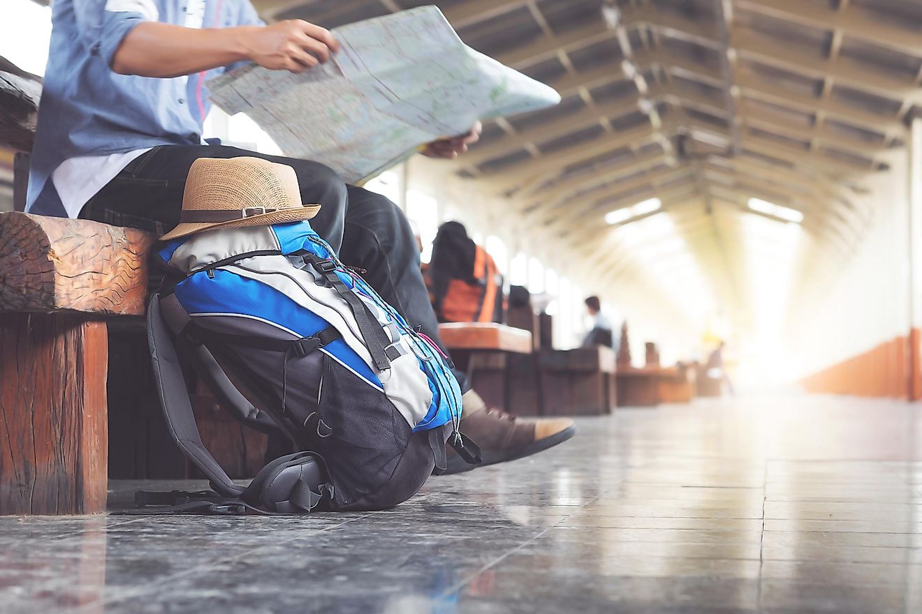 A traveler sits on a wooden bench, next to a backpack topped with a sun hat, inside of a long train station.