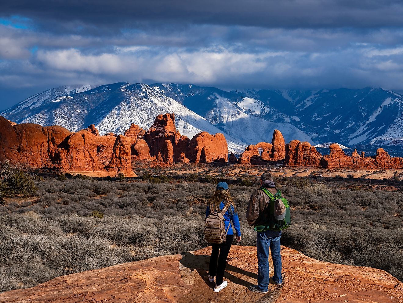 Couple enjoying a beautiful mountain view on a hiking trip in Utah, with the Windows Section of the park and snow-covered La Sal Mountains in the background. Arches National Park, Moab, Utah, USA.
