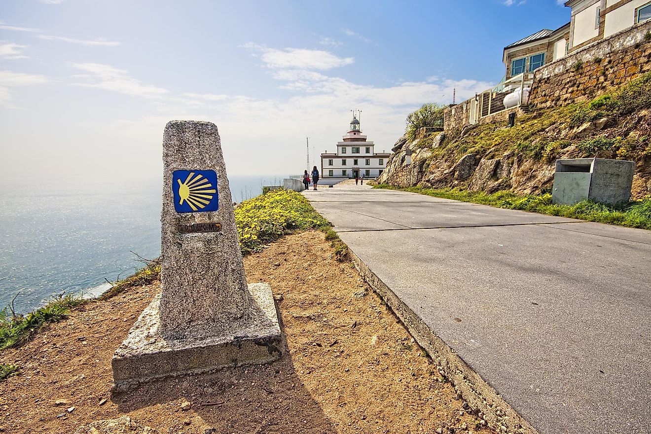 A stone pillar with a bright yellow and blue seashell logo and 0-kilometer indicator marks the end of the line for the Camino de Santiago. The lighthouse of Cape Finisterre and the vast Atlantic Ocean make up the background on a sunny afternoon.