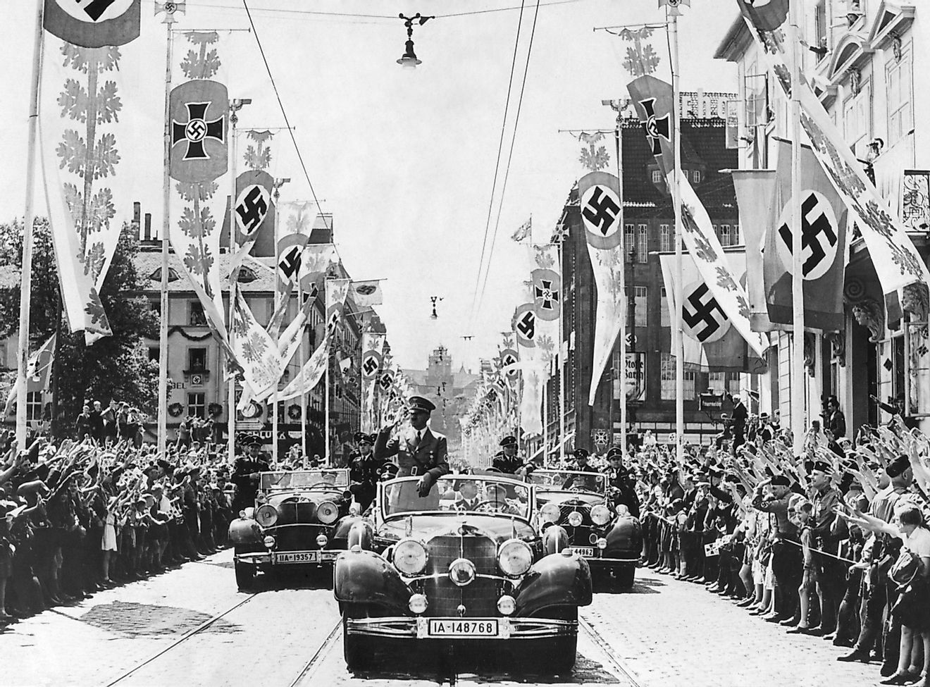 Adolf Hitler waving to crowds from his car at the head of a parade. The streets are decorated with various swastika banners.