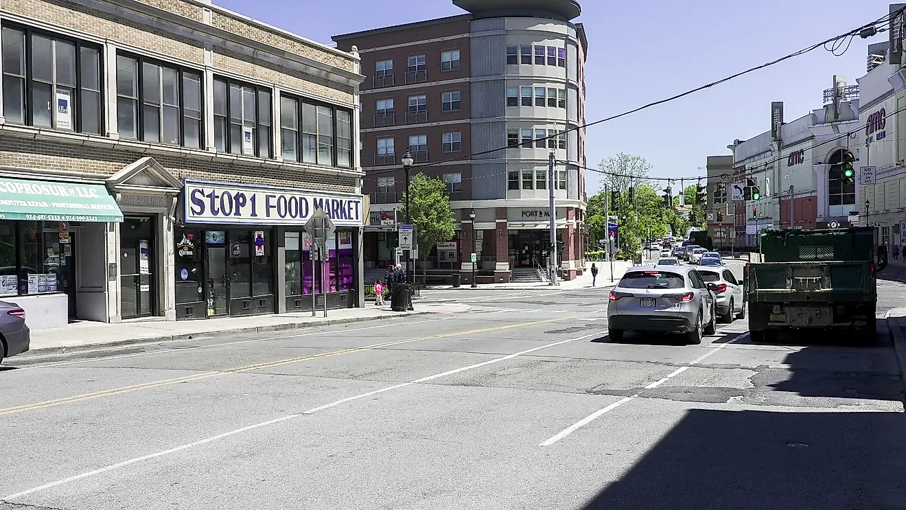Traffic and pedestrians moving past storefronts along a sunny urban street in Port Chester, New York. 