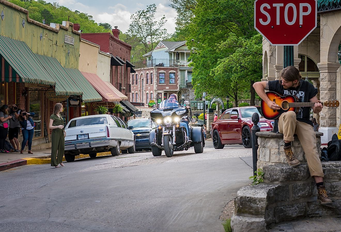 Biker visitors riding motorcycle downtown Eureka Springs, Man playing guitar at stop sign, freedom tourism on 2 wheels, vintage small american town. Image credit shuttersv via Shutterstock