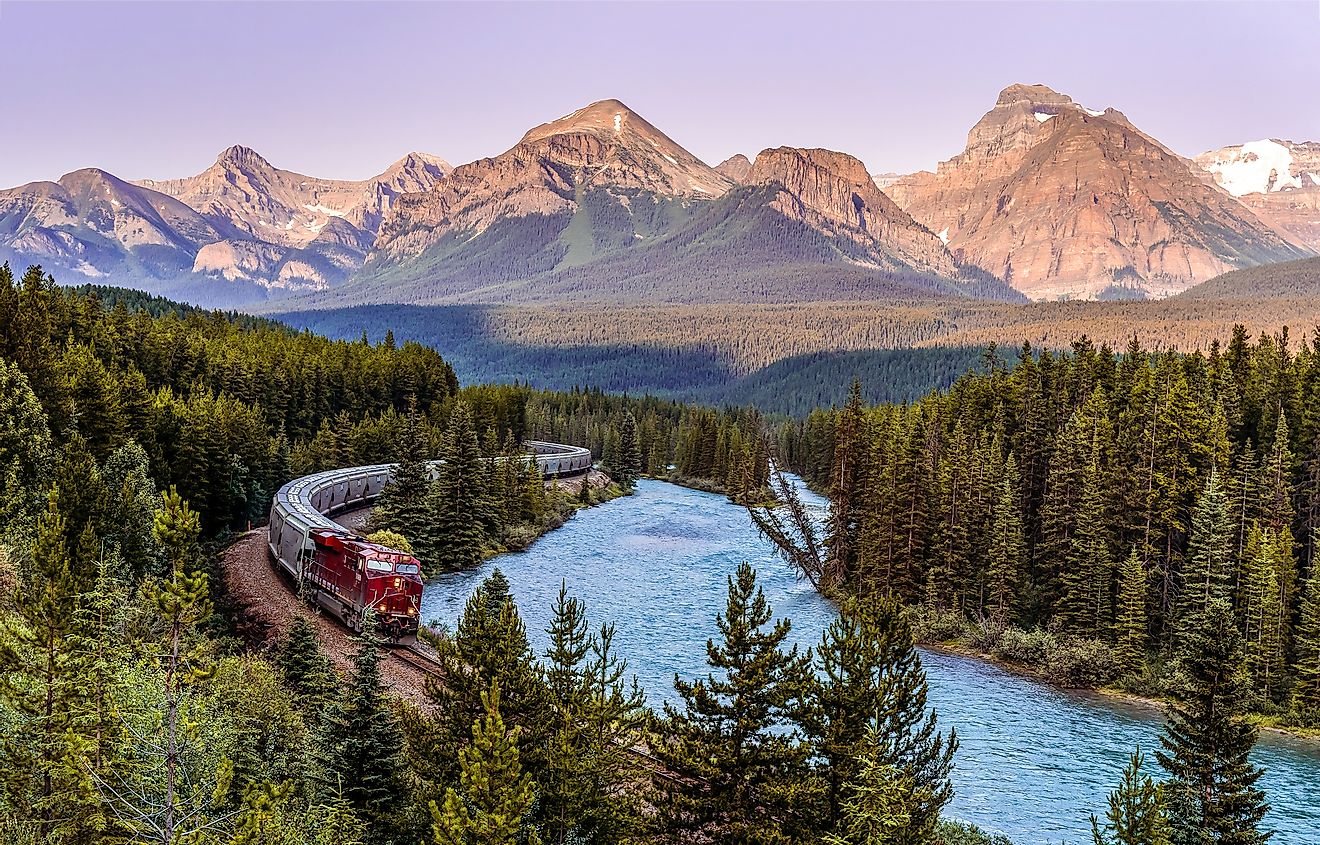 A passenger train winds through a forest, beside a river, with the European Alps standing tall in the background.