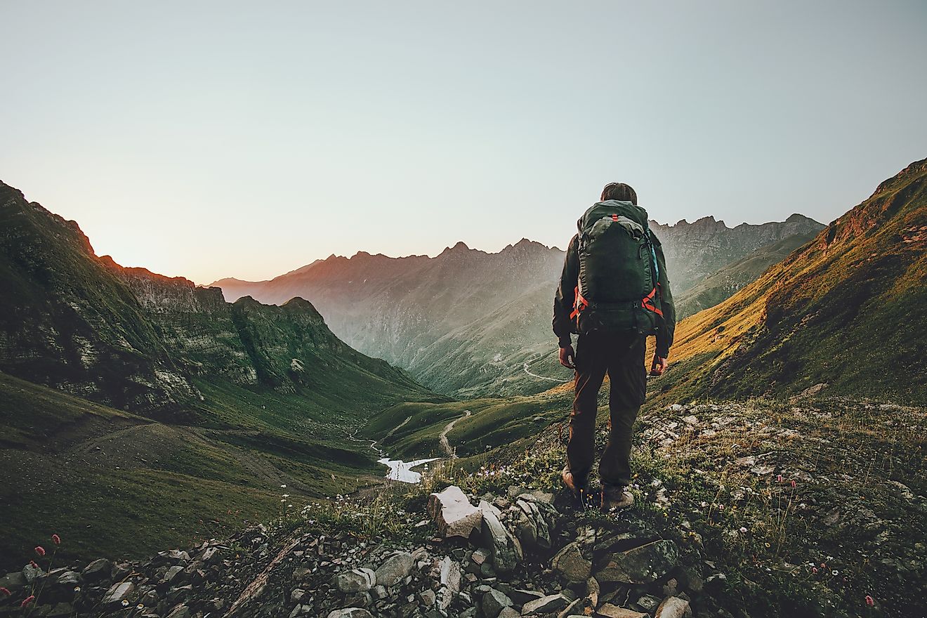 A backpacker stands above a green mountainous valley, looking back towards the setting sun.