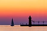 A vividly colorful twilight sky silhouettes a sailboat, people, and the lighthouse at South Haven, Michigan, on Lake Michigan.