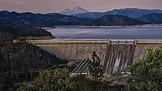 A picture of Shasta Dam with a lake and rocky mountains on the background during sunset. 