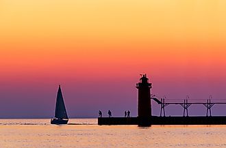 A vividly colorful twilight sky silhouettes a sailboat, people, and the lighthouse at South Haven, Michigan, on Lake Michigan.