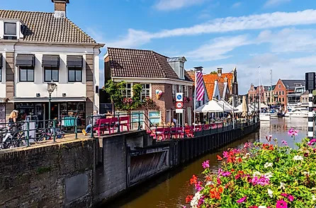 A vibrant canal-side scene in Lemmer, Netherlands, featuring historic Dutch buildings, a cozy café with pink benches, and colorful flowers.