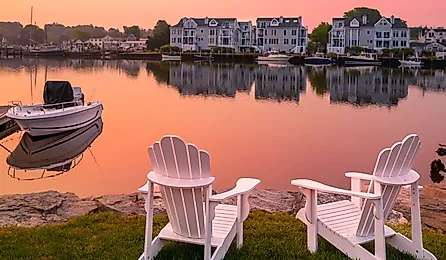 Sunrise, two white Adirondack chairs on the beach, a moored boat, and wildfire smoke over the Mystic River marina village in Connecticut.