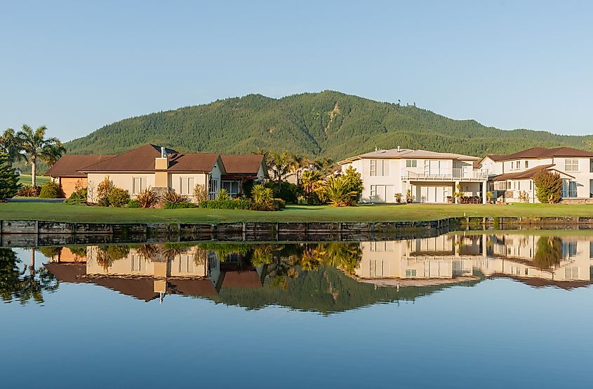 Idyllic residential home reflections in calm waterway foreground and green hill backdrop under blue sky at Pauanui New Zealand.