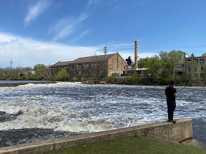 A man dressed in black balances on a concrete wall so he can fish in the wake at the base of a river dam.