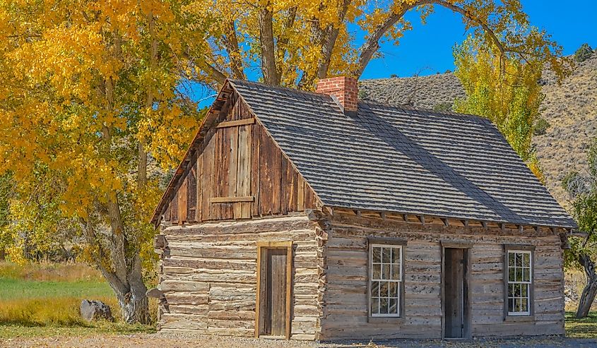 A rustic log cabin with a shingled roof sits under vibrant yellow autumn trees against a clear blue sky, conveying a peaceful, nostalgic ambiance.