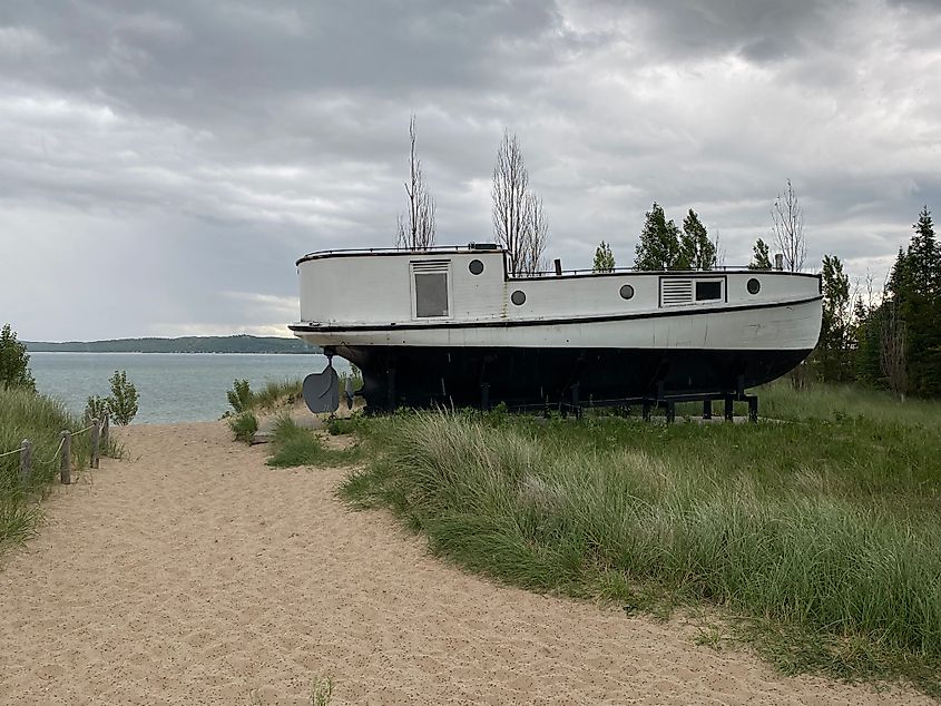A weathered white boat sits in a bed of wild grass next to a sandy trail and cove.