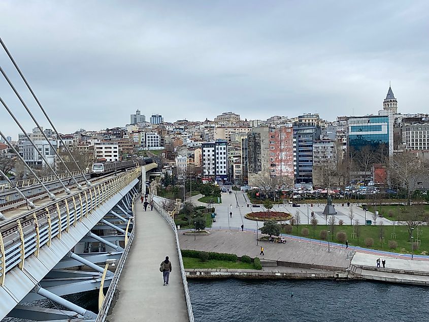 A river bridge with approaching train above and pedestrians below overlooks a river park and the weathered skyline of Istanbul.