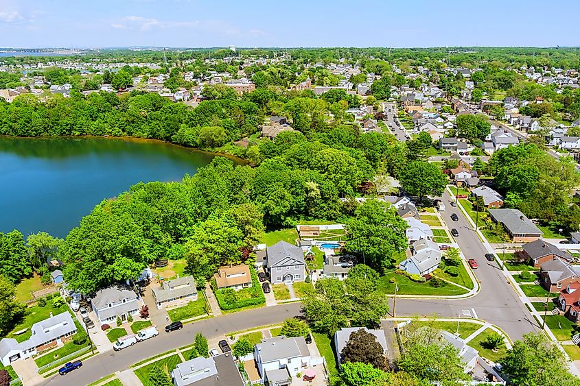 Aerial view of a suburban neighborhood in Sayreville, New Jersey, with single-family homes, tree-lined streets, and a large lake beside residential blocks on a clear day.
