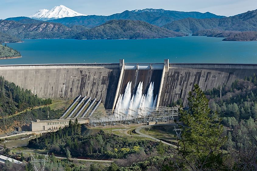 A picture of Shasta Dam surrounded by roads and trees with Shasta Lake and mountains on the background