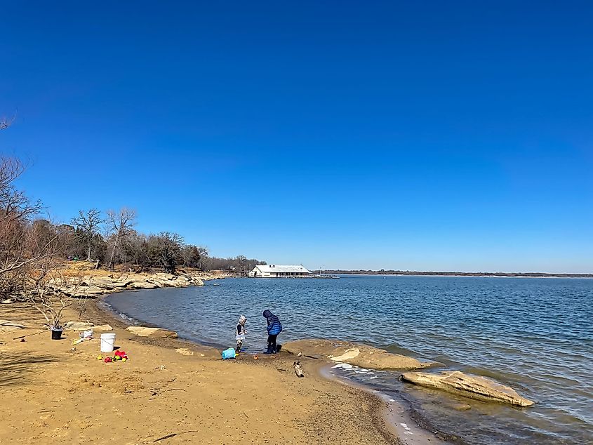 Unidentified children in winter jacket playing along the sandy rocky shoreline of Lake Lewisville during winter.