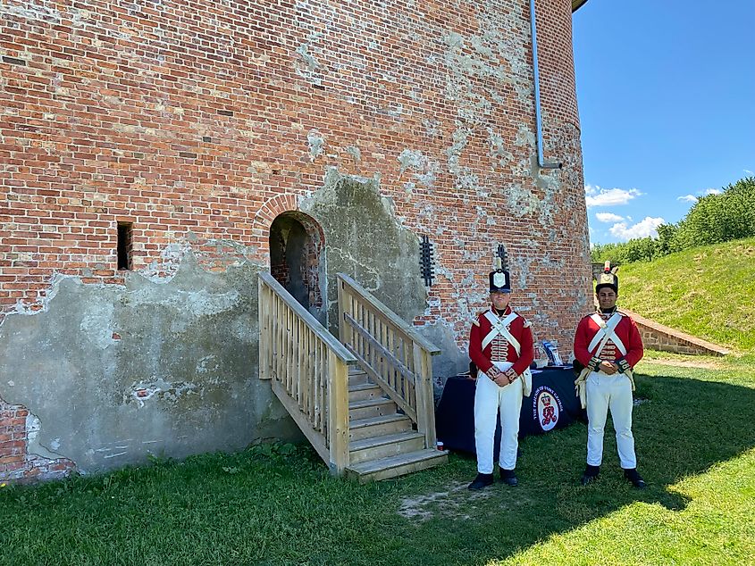 Two men dressed in red British military uniforms pose for a picture outside of a stone fort in a grassy field.