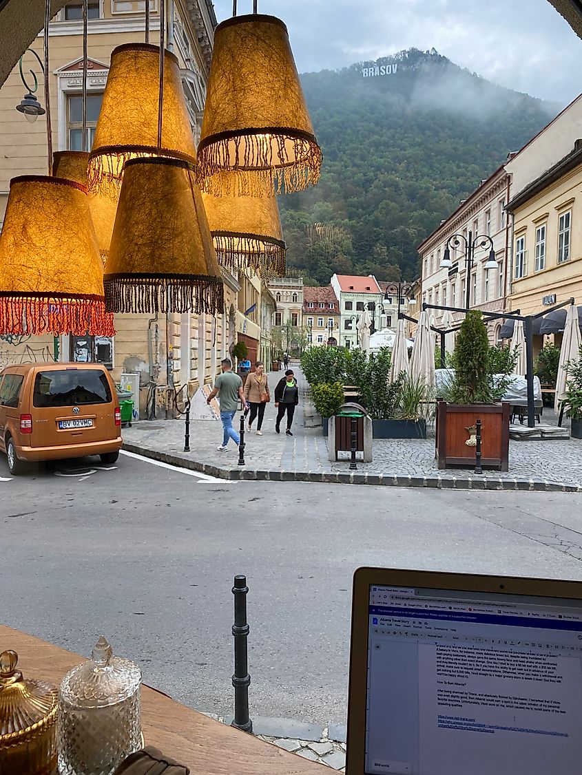 A laptop below old fashioned lamps, looking out a window onto the doings of an Eastern European mountain town. 
