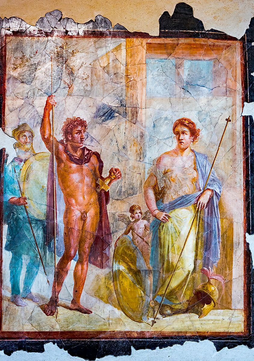 Fragmentary Roman wall painting showing seated figures, including Alexander the Great holding a sceptre beside a woman, with Eros nearby and a shield and helmet at the woman’s feet.