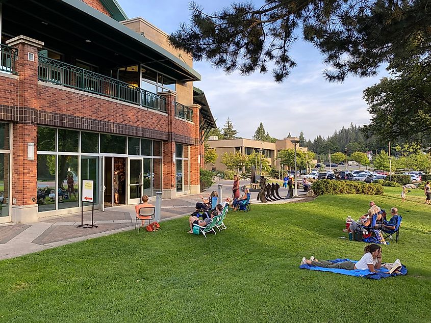 A small crowd sitting in a grassy park next to a large, red brick library. 