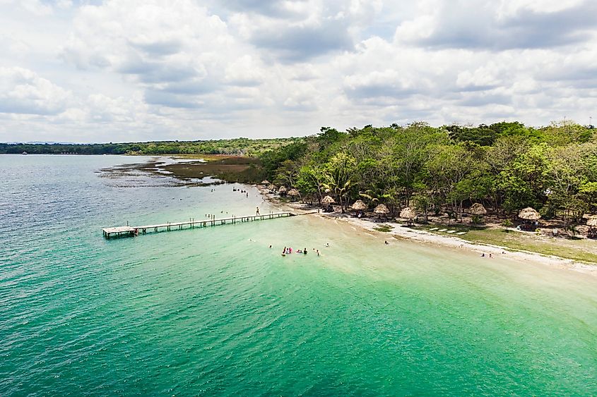 A wooden dock stretches out into sparkling blue waters with a sandy beach off to the side.